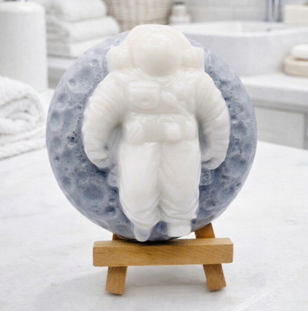 handmade vegan soap featuring a detailed astronaut sculpture floating against a detailed lunar surface background, displayed on a wooden easel.
