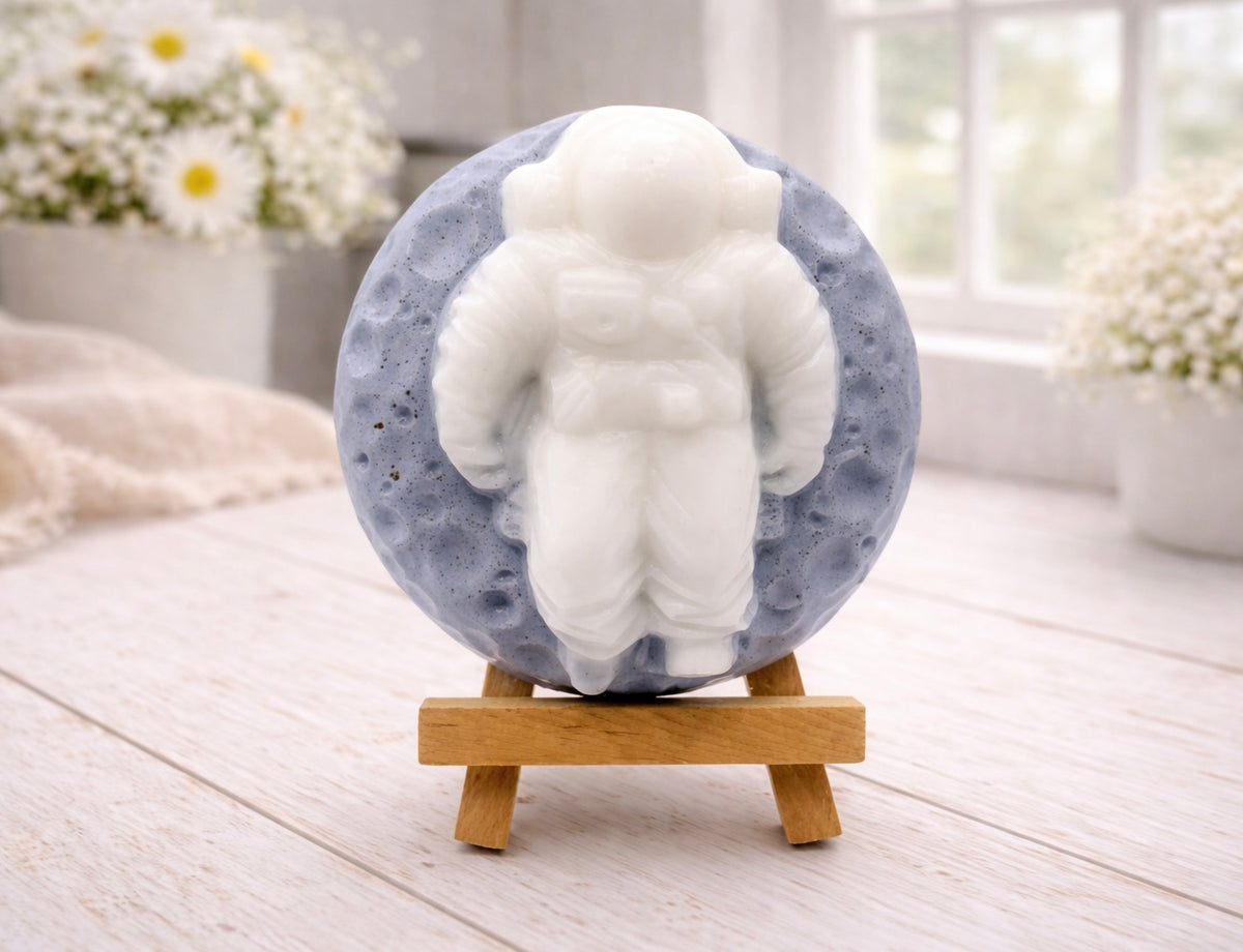 handmade vegan soap in the shape of a realistic astronaut sculpture sitting on the moon, featuring detailed spacesuit textures and craters on the lunar surface, displayed in a bright farmhouse decor setting.