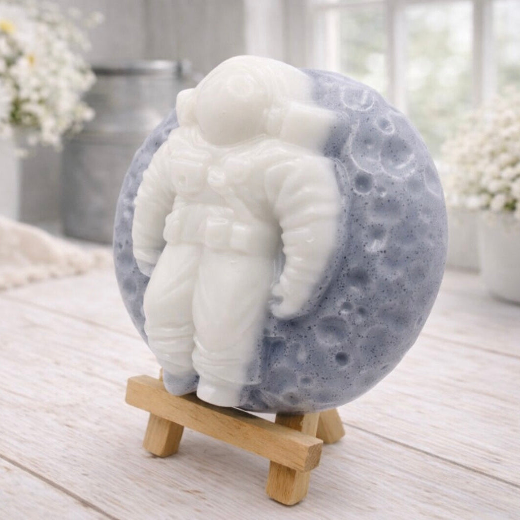 handmade vegan soap in the shape of a realistic astronaut sculpture sitting on the moon, featuring detailed spacesuit textures and craters on the lunar surface, displayed in a bright farmhouse decor setting.
