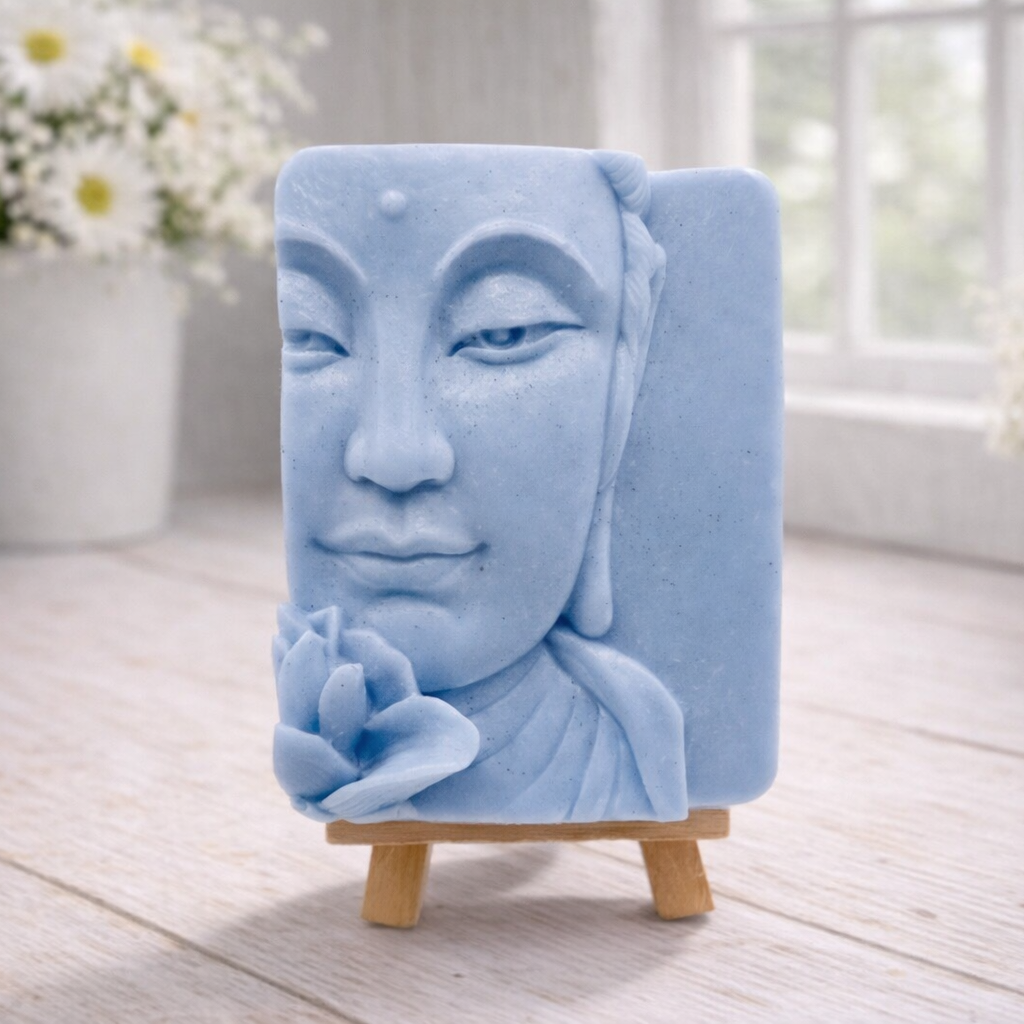 handmade vegan soap featuring a serene Buddha face relief with a lotus flower, displayed on a small wooden easel in a bright farmhouse decor setting with flowers and a rustic milk churn.