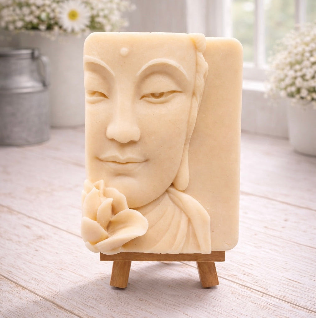 handmade vegan soap featuring a serene Buddha face relief with a lotus flower, displayed on a small wooden easel in a bright farmhouse decor setting with flowers and a rustic milk churn.