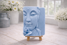 Load image into Gallery viewer, handmade vegan soap featuring a serene Buddha face relief with a lotus flower, displayed on a small wooden easel in a bright farmhouse decor setting with flowers and a rustic milk churn.
