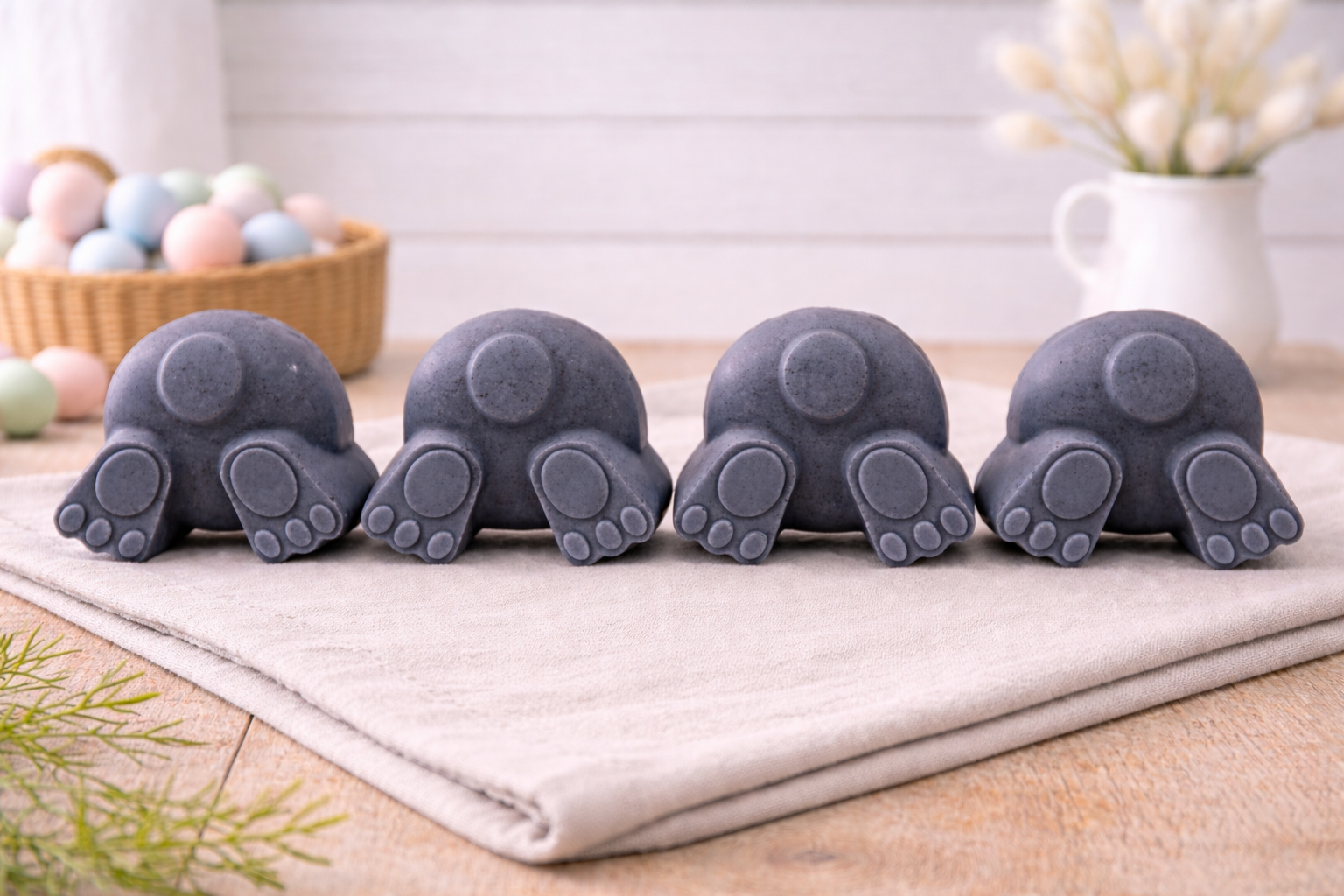 A set of four handmade vegan soaps in the shape of fluffy bunny tails and paws, displayed in a row on a neutral linen cloth with a basket of Easter eggs in the background.