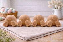 Load image into Gallery viewer, A set of four handmade vegan soaps in the shape of fluffy bunny tails and paws, displayed in a row on a neutral linen cloth with a basket of Easter eggs in the background.

