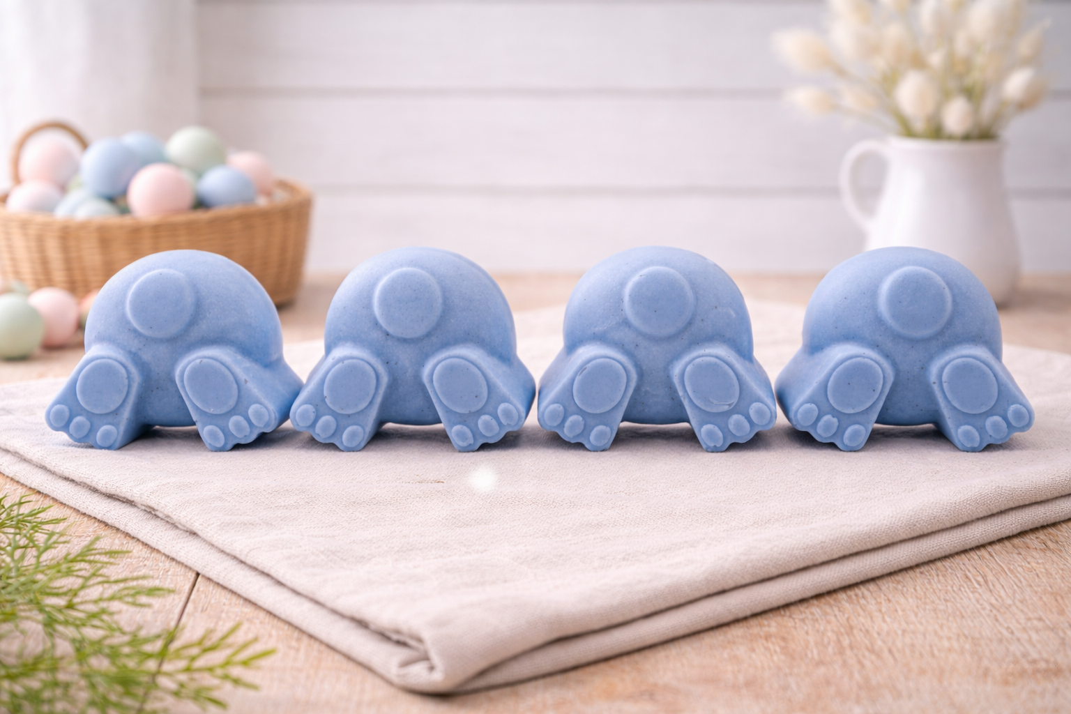 A set of four handmade vegan soaps in the shape of fluffy bunny tails and paws, displayed in a row on a neutral linen cloth with a basket of Easter eggs in the background.
