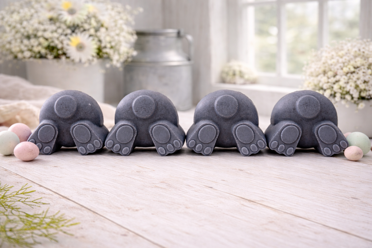 A set of four handmade vegan soaps in the shape of fluffy bunny tails and paws, displayed in a row on a neutral linen cloth with a basket of Easter eggs in the background.