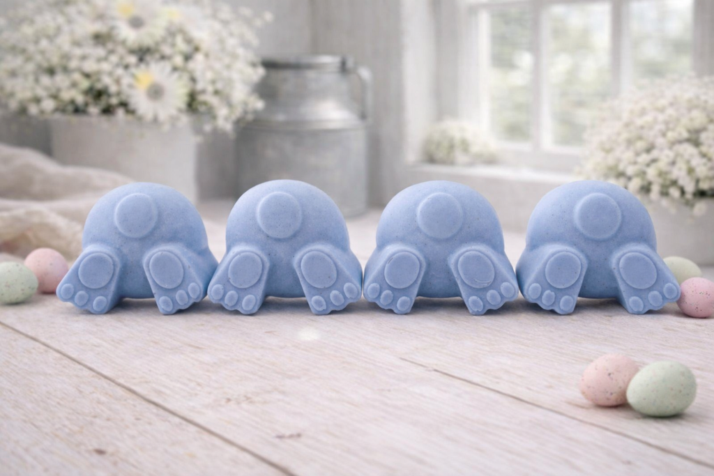 A set of four handmade vegan soaps in the shape of fluffy bunny tails and paws, displayed in a row on a neutral linen cloth with a basket of Easter eggs in the background.
