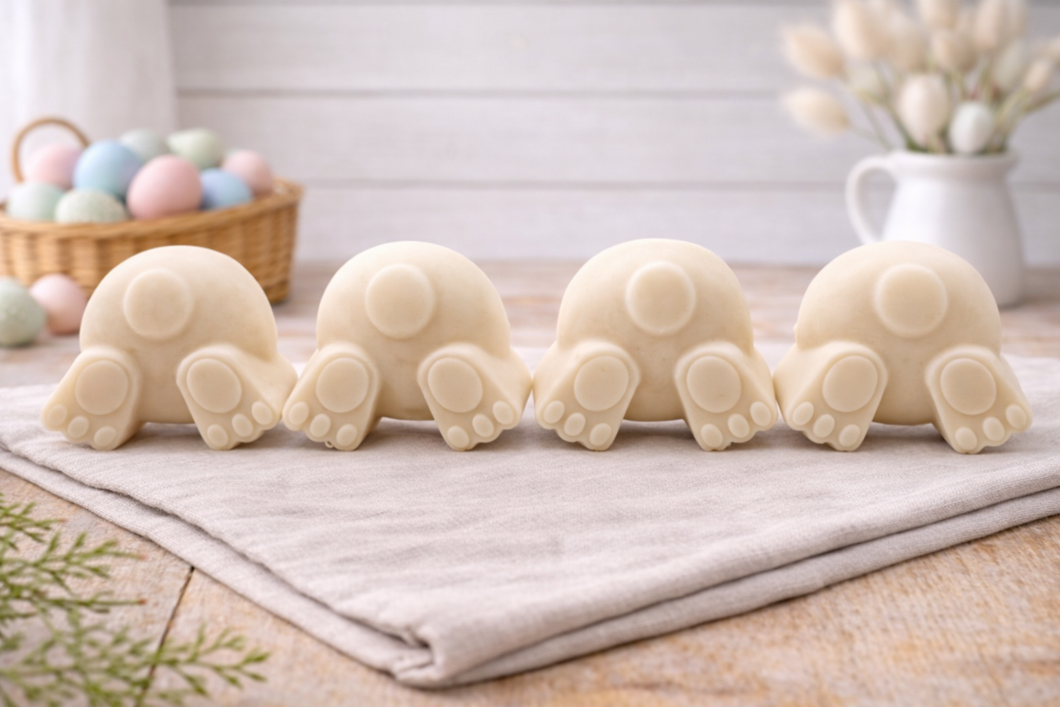 A set of four handmade vegan soaps in the shape of fluffy bunny tails and paws, displayed in a row on a neutral linen cloth with a basket of Easter eggs in the background.
