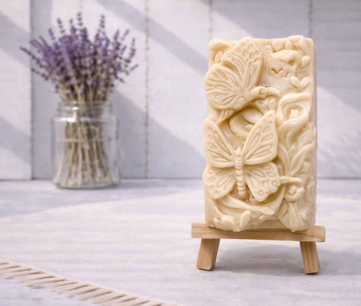 handmade vegan soap featuring a detailed 3D relief of butterflies and flowers, displayed on a wooden easel against a rustic farmhouse decor background with lavender.