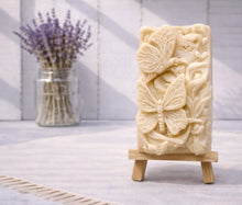 Load image into Gallery viewer, handmade vegan soap featuring a detailed 3D relief of butterflies and flowers, displayed on a wooden easel against a rustic farmhouse decor background with lavender.

