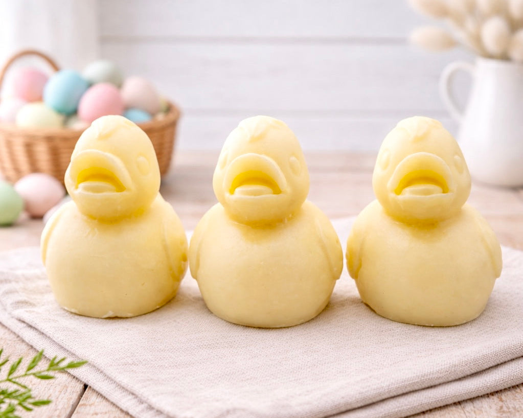 Three handmade vegan soaps in the shape of fluffy ducklings with open beaks, displayed on a neutral linen cloth in a bright farmhouse decor setting with Easter eggs in the background.