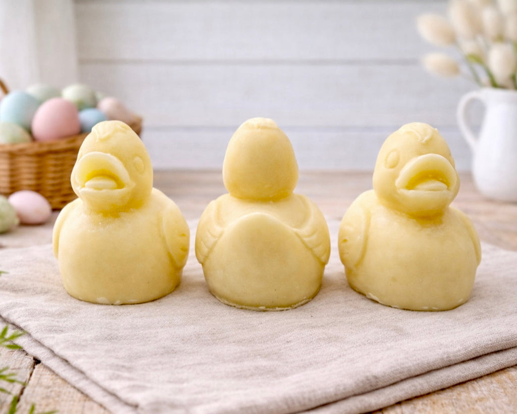 Three handmade vegan soaps in the shape of fluffy ducklings with open beaks, displayed on a neutral linen cloth in a bright farmhouse decor setting with Easter eggs in the background.