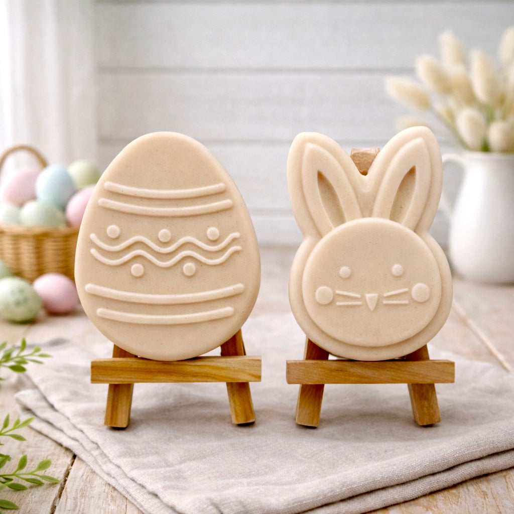 handmade vegan soap gift set featuring a cute bunny face and a decorated Easter egg with wave patterns, displayed on wooden easels against a rustic white farmhouse decor background with spring flowers.