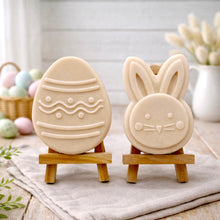 Load image into Gallery viewer, handmade vegan soap gift set featuring a cute bunny face and a decorated Easter egg with wave patterns, displayed on wooden easels against a rustic white farmhouse decor background with spring flowers.
