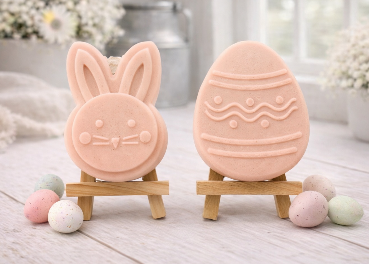 handmade vegan soap gift set featuring a cute bunny face and a decorated Easter egg with wave patterns, displayed on wooden easels against a rustic white farmhouse decor background with spring flowers.