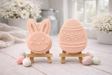 Load image into Gallery viewer, handmade vegan soap gift set featuring a cute bunny face and a decorated Easter egg with wave patterns, displayed on wooden easels against a rustic white farmhouse decor background with spring flowers.
