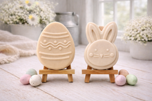 Load image into Gallery viewer, handmade vegan soap gift set featuring a cute bunny face and a decorated Easter egg with wave patterns, displayed on wooden easels against a rustic white farmhouse decor background with spring flowers.
