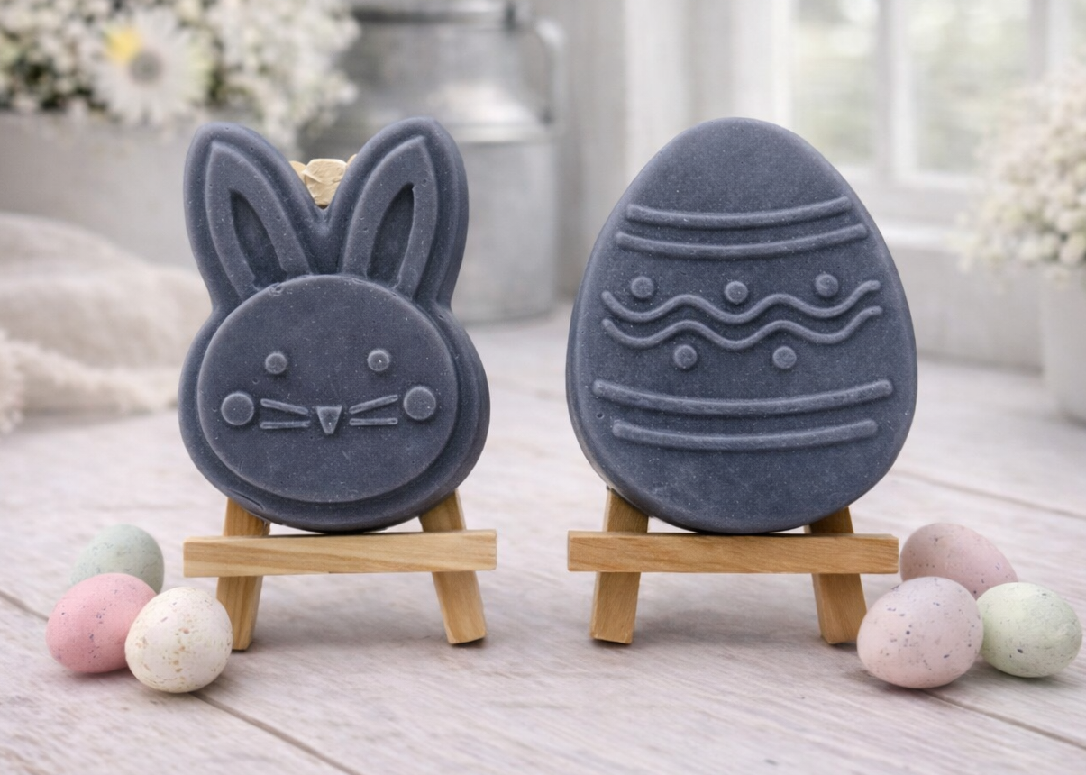 handmade vegan soap gift set featuring a cute bunny face and a decorated Easter egg with wave patterns, displayed on wooden easels against a rustic white farmhouse decor background with spring flowers.