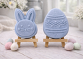 handmade vegan soap gift set featuring a cute bunny face and a decorated Easter egg with wave patterns, displayed on wooden easels against a rustic white farmhouse decor background with spring flowers.