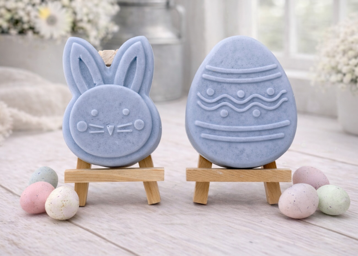 handmade vegan soap gift set featuring a cute bunny face and a decorated Easter egg with wave patterns, displayed on wooden easels against a rustic white farmhouse decor background with spring flowers.