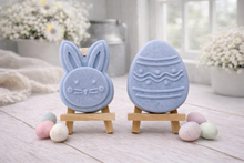 Load image into Gallery viewer, handmade vegan soap gift set featuring a cute bunny face and a decorated Easter egg with wave patterns, displayed on wooden easels against a rustic white farmhouse decor background with spring flowers.
