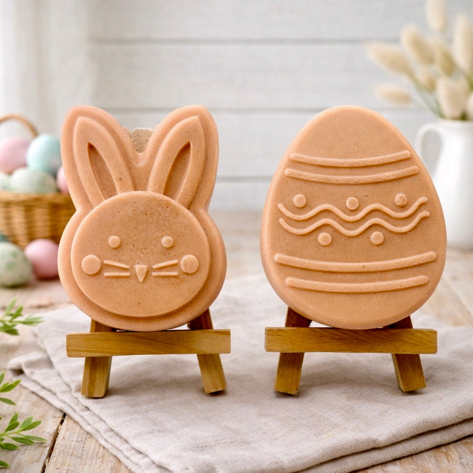 handmade vegan soap gift set featuring a cute bunny face and a decorated Easter egg with wave patterns, displayed on wooden easels against a rustic white farmhouse decor background with spring flowers.