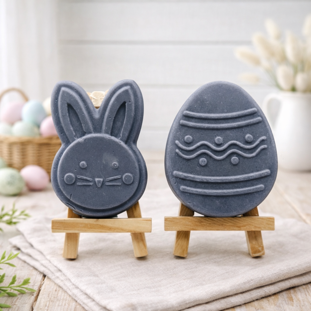 handmade vegan soap gift set featuring a cute bunny face and a decorated Easter egg with wave patterns, displayed on wooden easels against a rustic white farmhouse decor background with spring flowers.