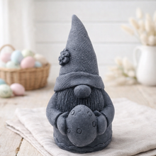 Load image into Gallery viewer, handmade vegan soap in the shape of a sitting gnome holding a decorated Easter egg, featuring a tall pointed hat with a flower detail and a textured beard, displayed on a neutral cloth in a bright spring setting.
