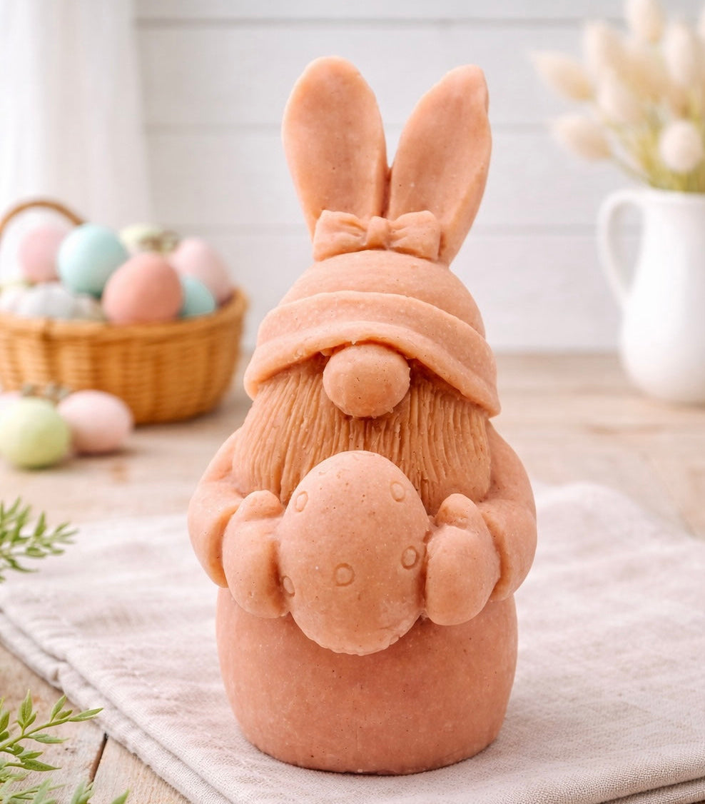 handmade vegan soap sculpture of a gnome wearing a rabbit-ear hat with a bow, holding a decorated Easter egg, set against a rustic background with a basket of pastel eggs.