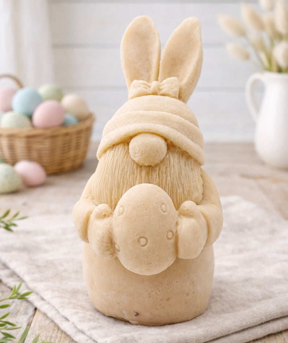 handmade vegan soap sculpture of a gnome wearing a rabbit-ear hat with a bow, holding a decorated Easter egg, set against a rustic background with a basket of pastel eggs.