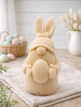 Load image into Gallery viewer, handmade vegan soap sculpture of a gnome wearing a rabbit-ear hat with a bow, holding a decorated Easter egg, set against a rustic background with a basket of pastel eggs.
