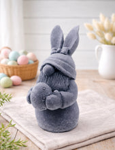 Load image into Gallery viewer, handmade vegan soap sculpture of a gnome wearing a rabbit-ear hat with a bow, holding a decorated Easter egg, set against a rustic background with a basket of pastel eggs.
