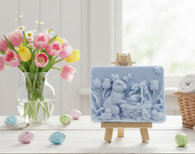 Load image into Gallery viewer, handmade vegan soap featuring a highly detailed 3D relief of a rabbit in a garden with tulips and an Easter basket, displayed on a wooden easel in a rustic farmhouse decor setting.
