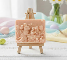 Load image into Gallery viewer, handmade vegan soap featuring a highly detailed 3D relief of a rabbit in a garden with tulips and an Easter basket, displayed on a wooden easel in a rustic farmhouse decor setting.
