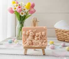 Load image into Gallery viewer, handmade vegan soap featuring a highly detailed 3D relief of a rabbit in a garden with tulips and an Easter basket, displayed on a wooden easel in a rustic farmhouse decor setting.
