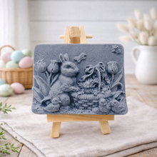 Load image into Gallery viewer, handmade vegan soap featuring a highly detailed 3D relief of a rabbit in a garden with tulips and an Easter basket, displayed on a wooden easel in a rustic farmhouse decor setting.
