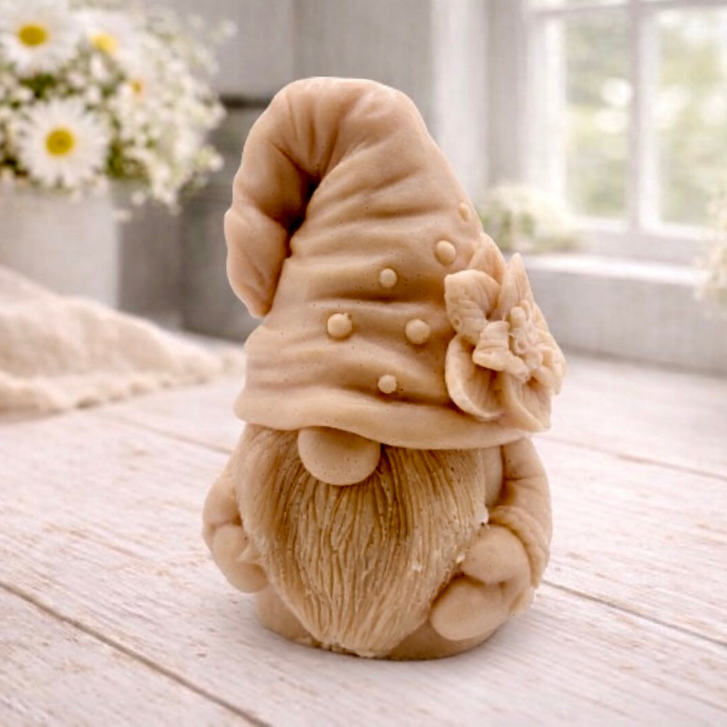 handmade vegan soap in the shape of a garden gnome sculpture with a long beard and a tall hat adorned with a large rose, displayed on a rustic white wooden surface in a bright farmhouse decor setting.