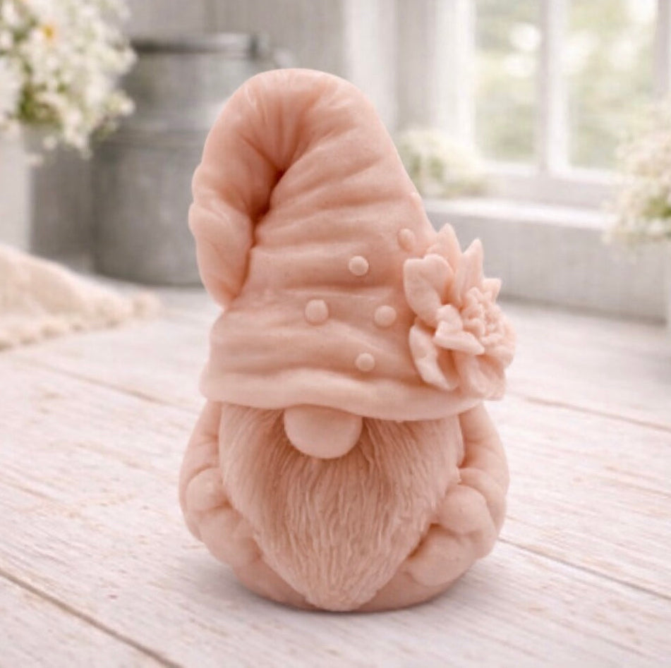 handmade vegan soap in the shape of a garden gnome sculpture with a long beard and a tall hat adorned with a large rose, displayed on a rustic white wooden surface in a bright farmhouse decor setting.
