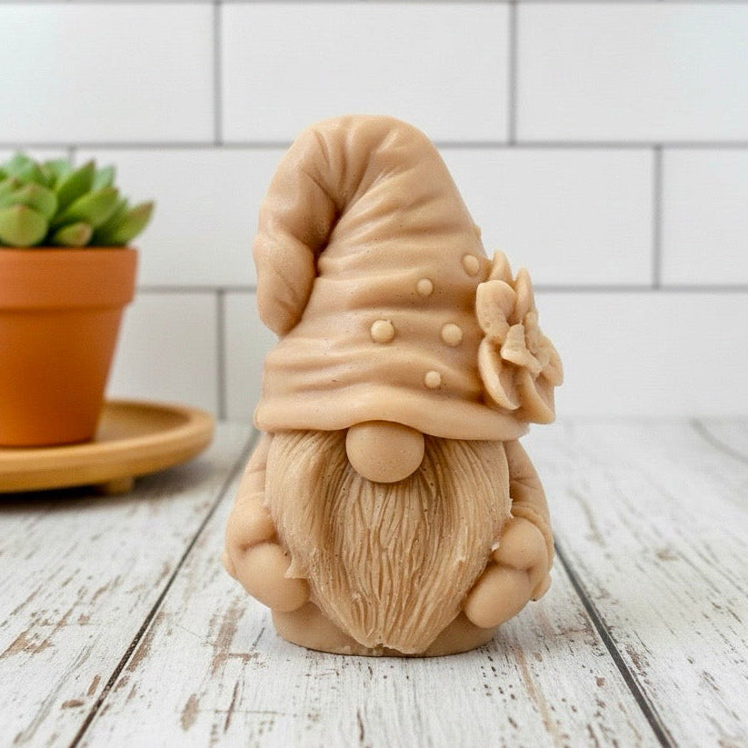 handmade vegan soap in the shape of a garden gnome sculpture with a long beard and a tall hat adorned with a large rose, displayed on a rustic white wooden surface in a bright farmhouse decor setting.