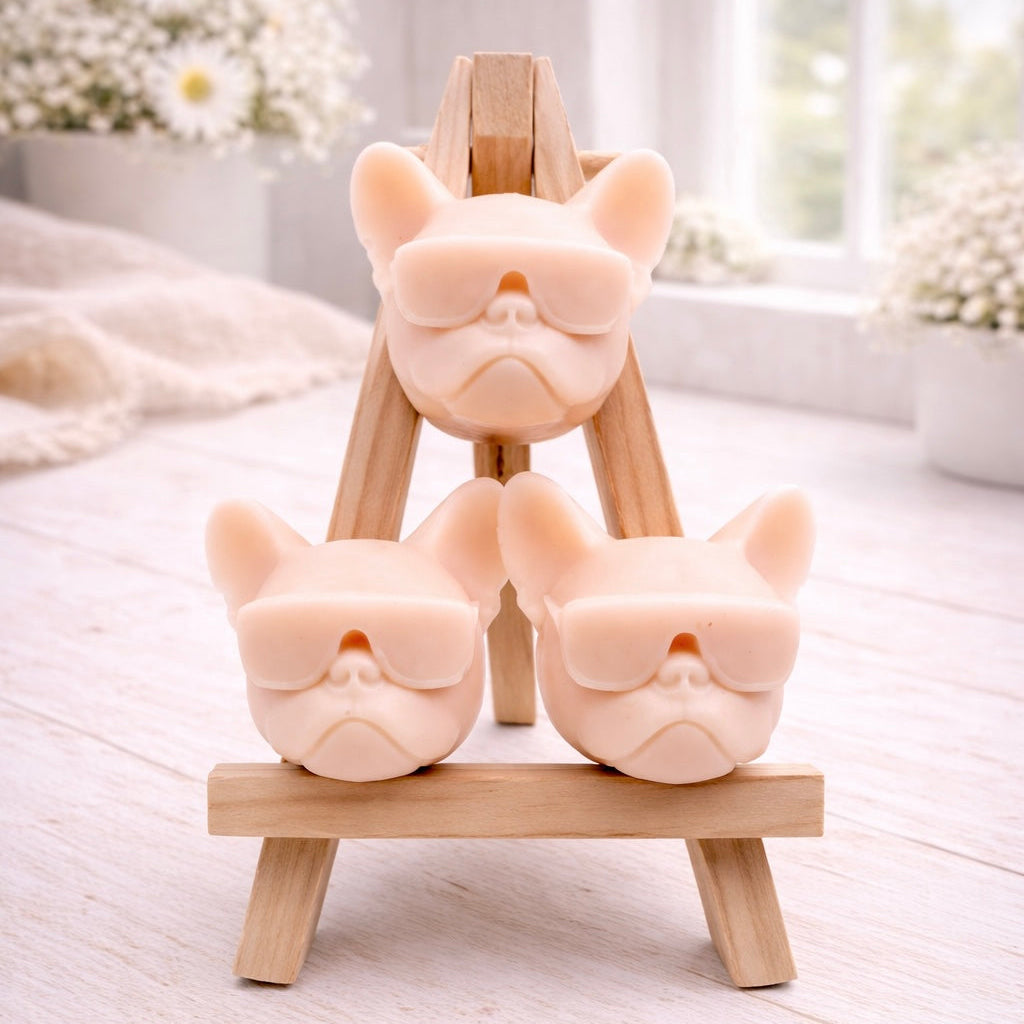 A set of three handmade vegan soaps in the shape of French Bulldog heads wearing sunglasses, displayed on a wooden easel in a bright, modern bathroom setting.