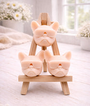 Load image into Gallery viewer, A set of three handmade vegan soaps in the shape of French Bulldog heads wearing sunglasses, displayed on a wooden easel in a bright, modern bathroom setting.
