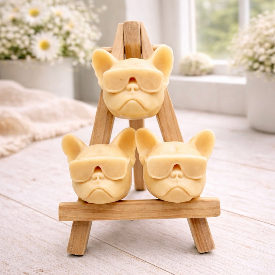 A set of three handmade vegan soaps in the shape of French Bulldog heads wearing sunglasses, displayed on a wooden easel in a bright, modern bathroom setting.