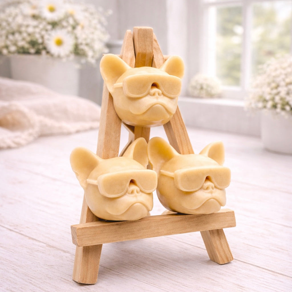 A set of three handmade vegan soaps in the shape of French Bulldog heads wearing sunglasses, displayed on a wooden easel in a bright, modern bathroom setting.