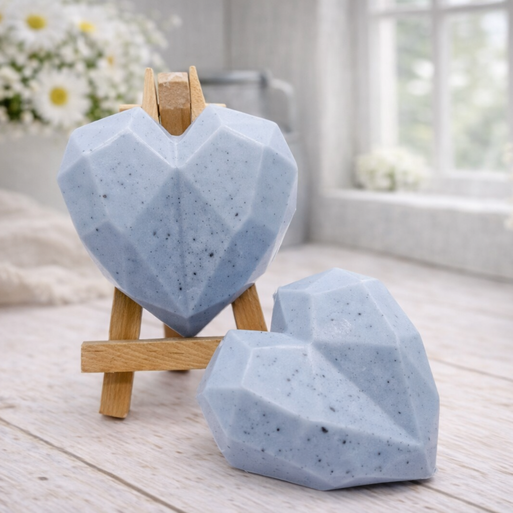 Two handmade vegan soaps in a contemporary geometric heart shape, with one displayed on a small wooden easel, set against a bright farmhouse decor background with white flowers.