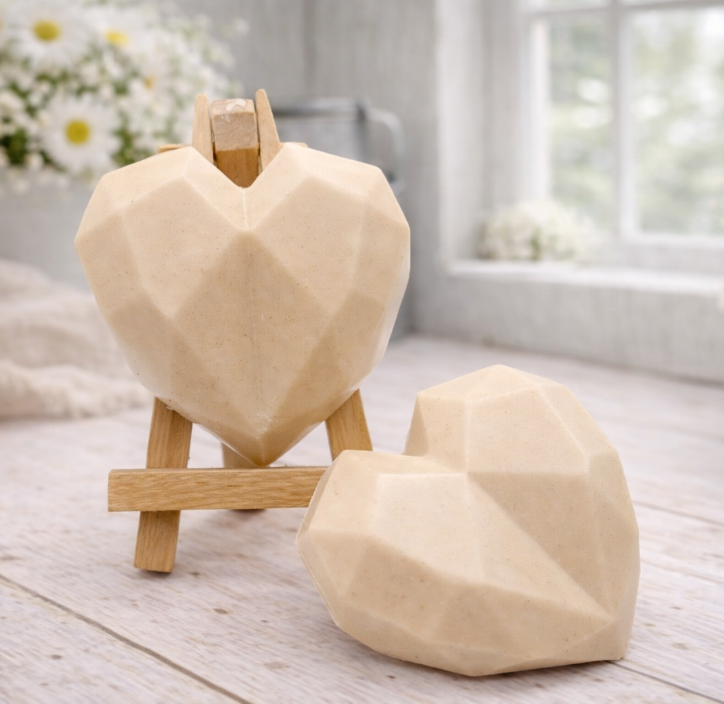 Two handmade vegan soaps in a contemporary geometric heart shape, with one displayed on a small wooden easel, set against a bright farmhouse decor background with white flowers.