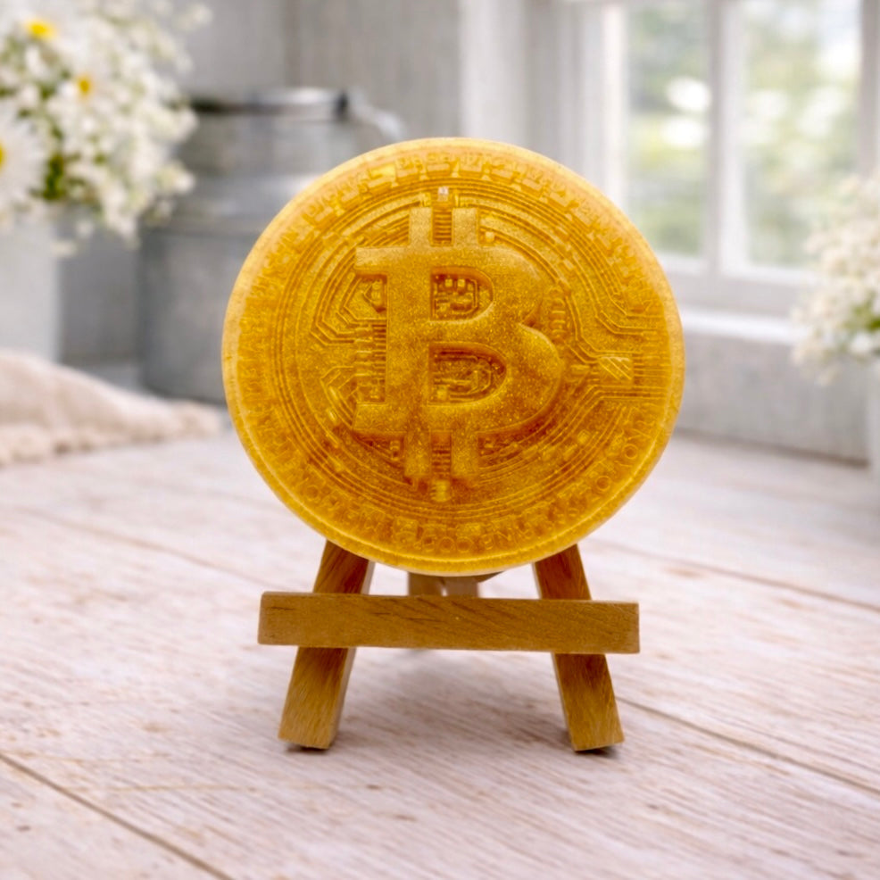 handmade vegan soap in the shape of a Bitcoin coin with a shimmering golden finish, featuring a detailed relief of the "B" symbol and circuit board patterns, displayed on a small wooden easel in a bright farmhouse decor setting.