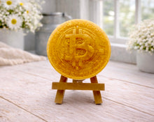 Load image into Gallery viewer, handmade vegan soap in the shape of a Bitcoin coin with a shimmering golden finish, featuring a detailed relief of the "B" symbol and circuit board patterns, displayed on a small wooden easel in a bright farmhouse decor setting.

