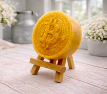 Load image into Gallery viewer, handmade vegan soap in the shape of a Bitcoin coin with a shimmering golden finish, featuring a detailed relief of the "B" symbol and circuit board patterns, displayed on a small wooden easel in a bright farmhouse decor setting.
