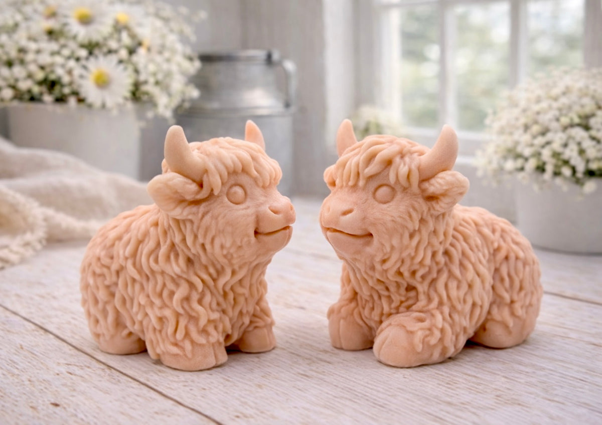 A highly detailed handmade vegan soap sculpted in the shape of a highland cow with long, flowing fur texture, a sweet face with a large snout, and prominent horns, displayed on a light wooden surface in a bright farmhouse setting.