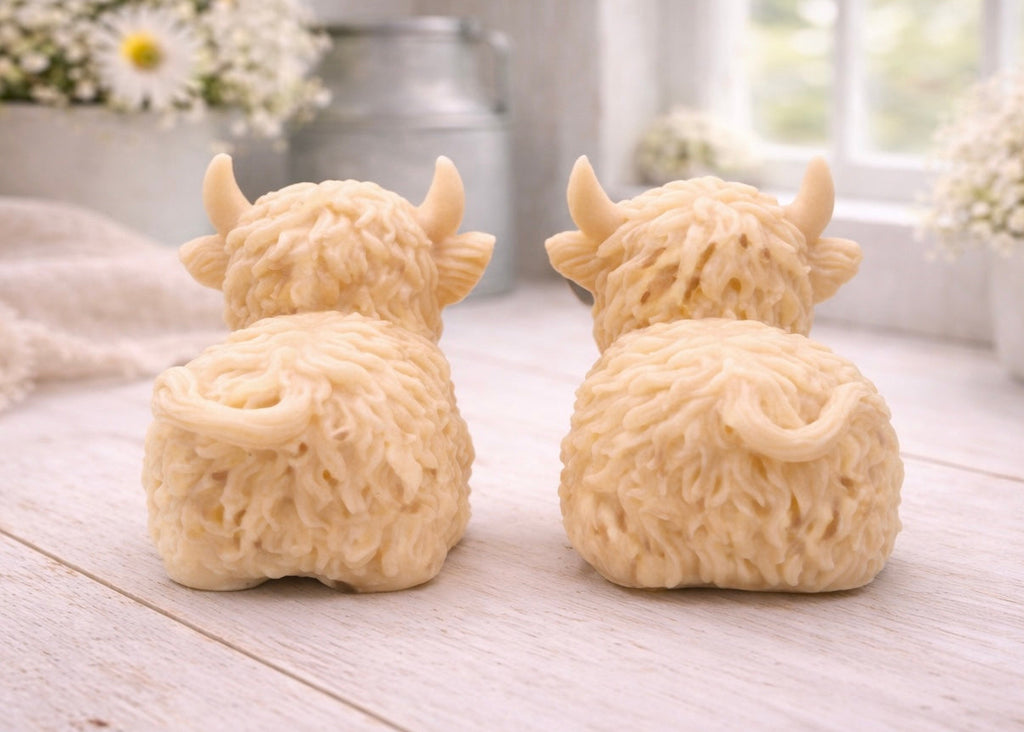 A highly detailed handmade vegan soap sculpted in the shape of a highland cow with long, flowing fur texture, a sweet face with a large snout, and prominent horns, displayed on a light wooden surface in a bright farmhouse setting.